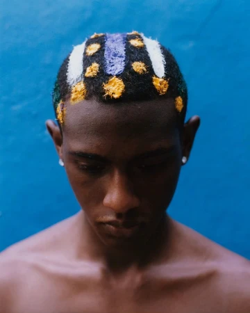 Close-up of a young adult with dark skin, adorned with white, purple, and yellow designs on a short haircut, set against a blue background.
