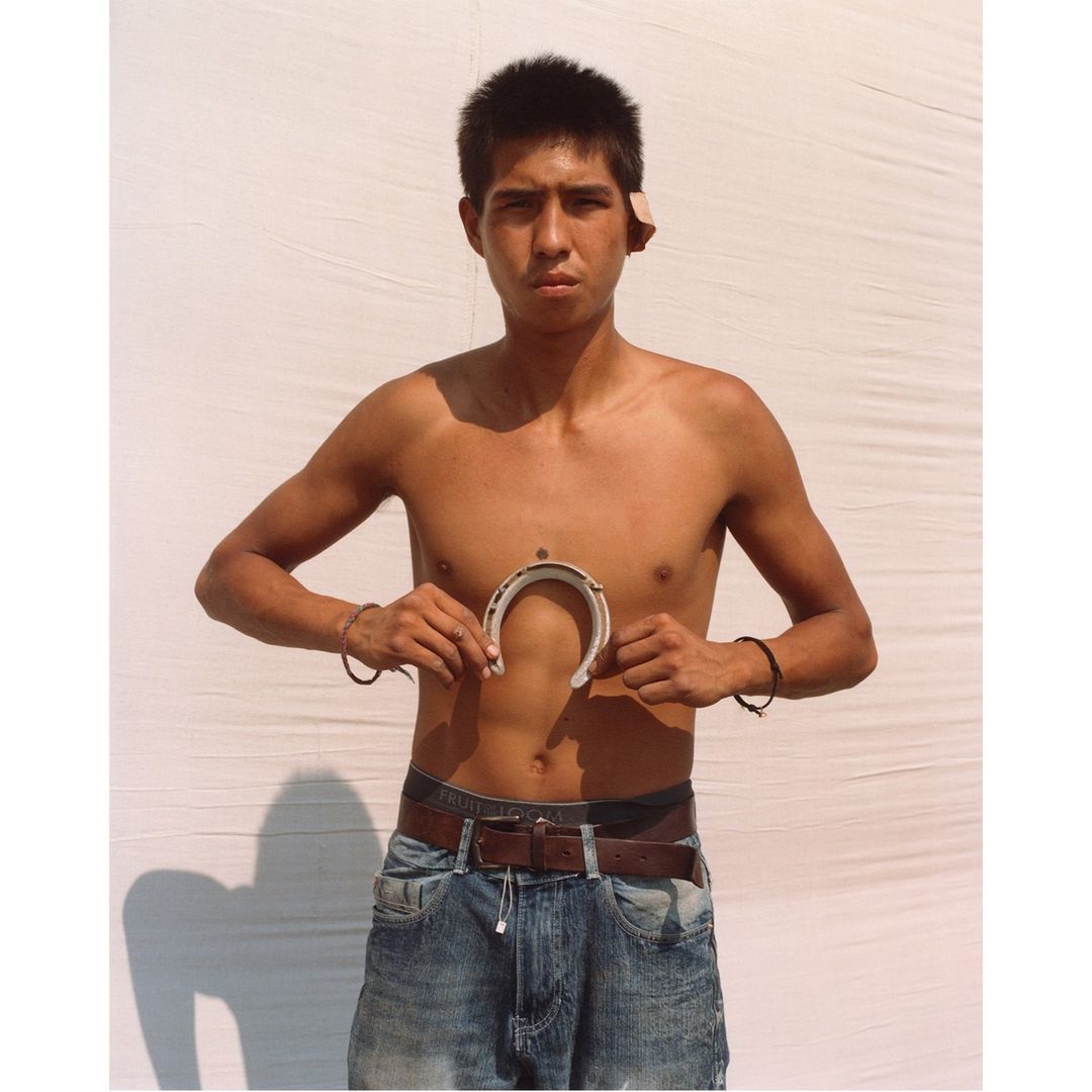 Young adult male holds a horseshoe in front of white backdrop, wearing jeans and bracelets, with a serious expression and direct gaze.