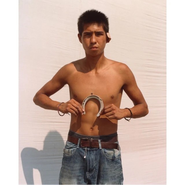 Young adult male holds a horseshoe in front of white backdrop, wearing jeans and bracelets, with a serious expression and direct gaze.