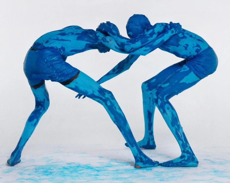 Two figures covered in blue paint grapple in a dynamic studio shot, showcasing body art and performance in a minimalist setting.