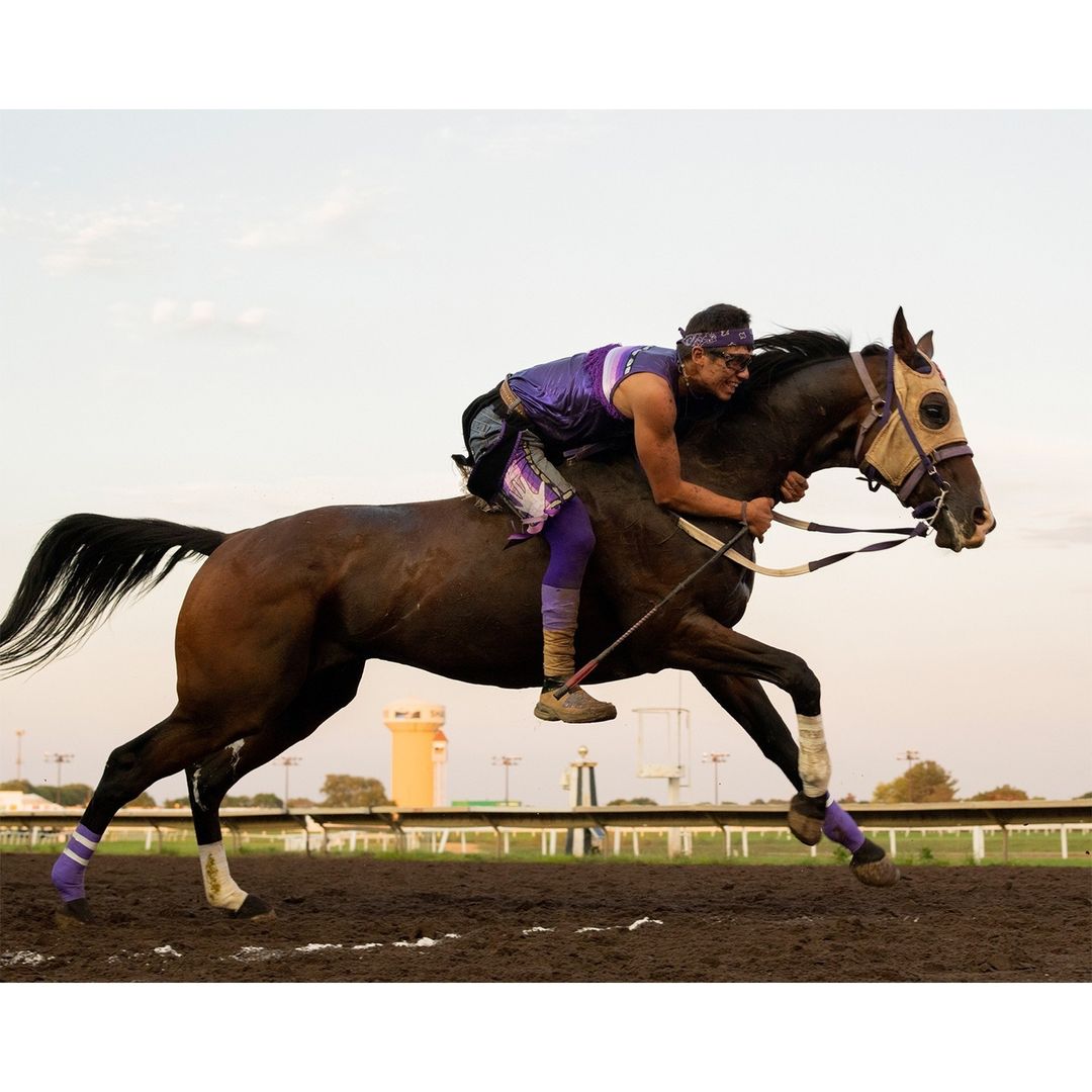 Horse race featuring a rider wearing purple, moving at full speed on a dirt track with a building and sky in the background.