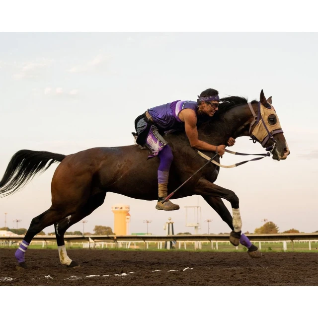 Horse race featuring a rider wearing purple, moving at full speed on a dirt track with a building and sky in the background.