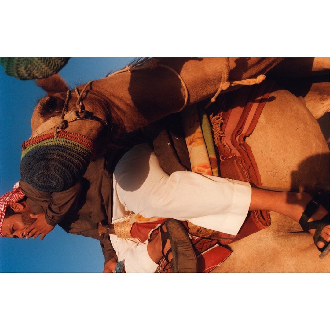 Man riding a camel with blue sky in the background. He is wearing a traditional headscarf and white clothing, sitting in an open desert area.