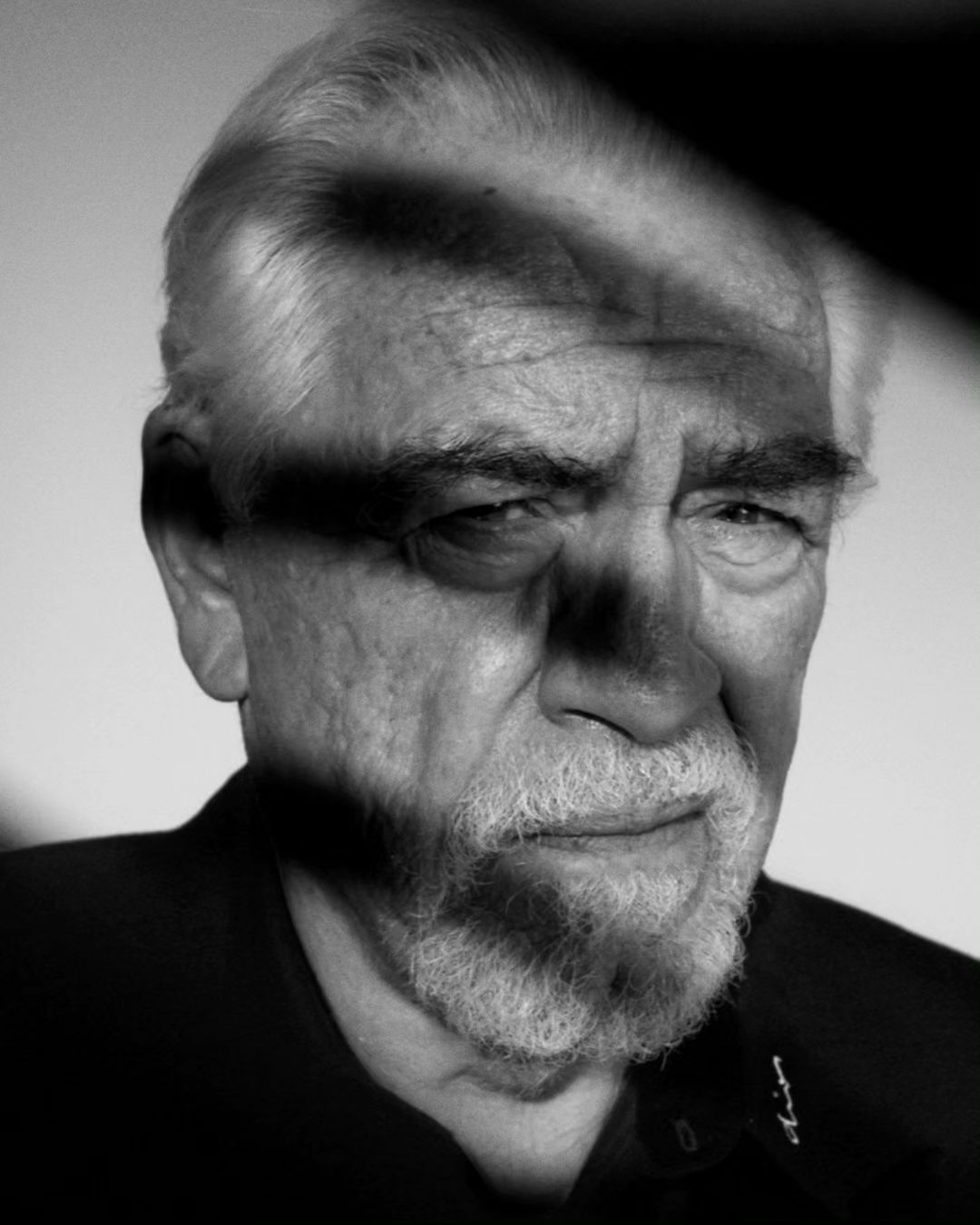 Monochrome portrait of a mature adult male with a white beard, photographed indoors with partial shadow casting across the face.
