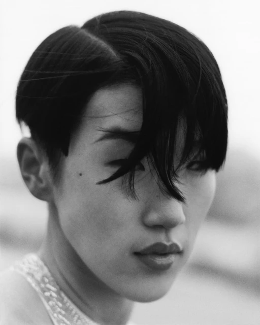 Monochrome portrait of an East Asian adult female with short dark hair, bold eye makeup, and glossy lips against a blurred background.