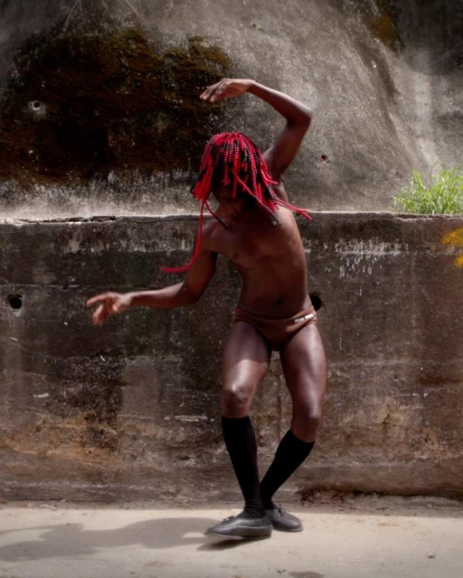 Adult male with red braided hair dances against a distressed concrete wall in athletic briefs, black socks, and leather shoes.