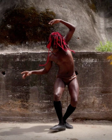 Adult male with red braided hair dances against a distressed concrete wall in athletic briefs, black socks, and leather shoes.