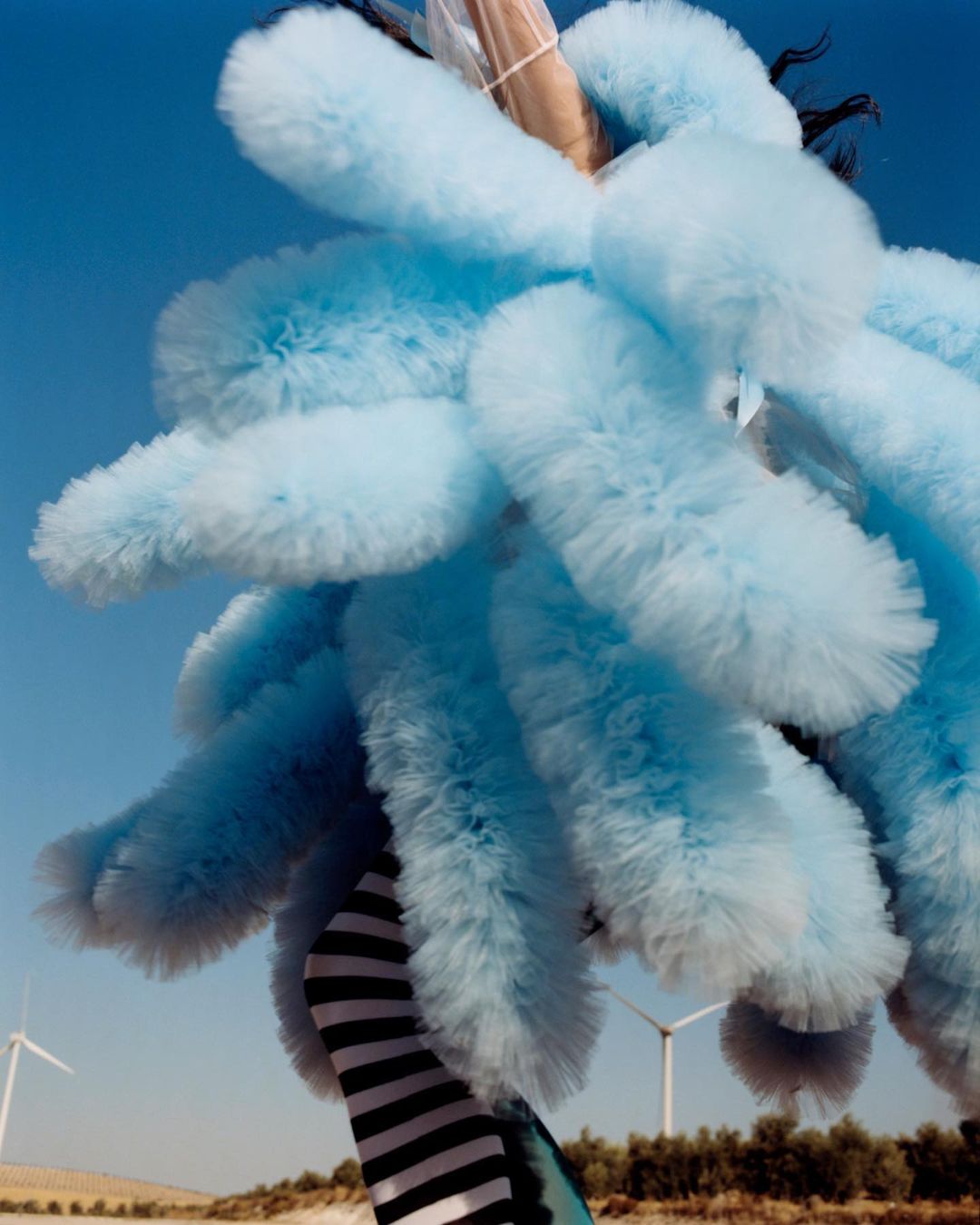 A person in a blue, fluffy costume poses outdoors, with wind turbines visible in the background under a clear blue sky.