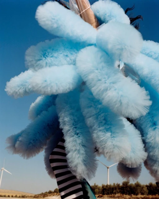 A person in a blue, fluffy costume poses outdoors, with wind turbines visible in the background under a clear blue sky.