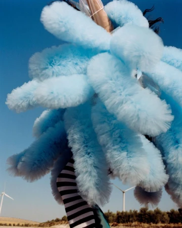 A person in a blue, fluffy costume poses outdoors, with wind turbines visible in the background under a clear blue sky.