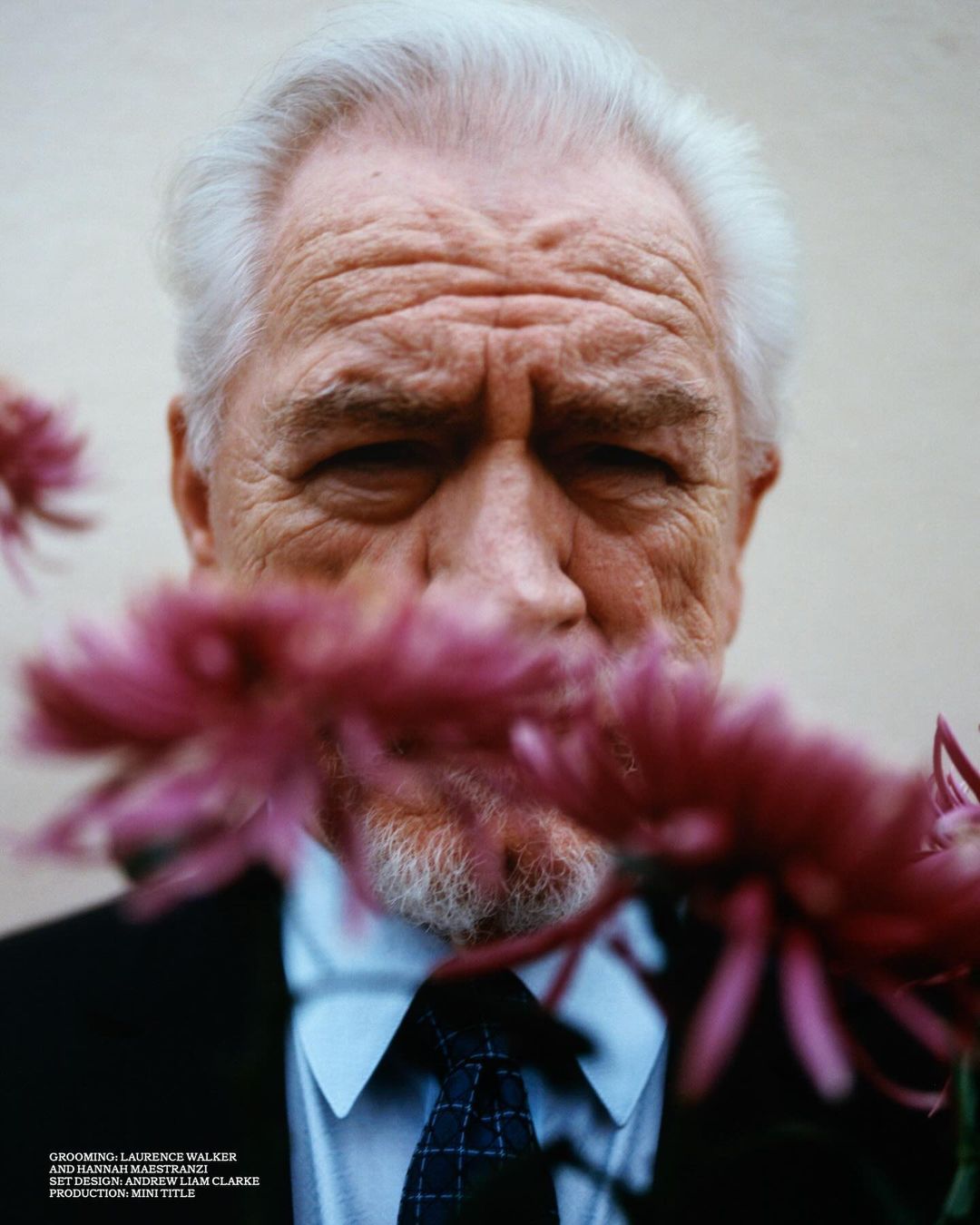 Portrait of an elderly adult man with white hair, wearing a suit and tie, partially obscured by pink flowers against a plain background.