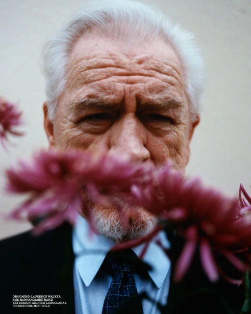 Portrait of an elderly adult man with white hair, wearing a suit and tie, partially obscured by pink flowers against a plain background.