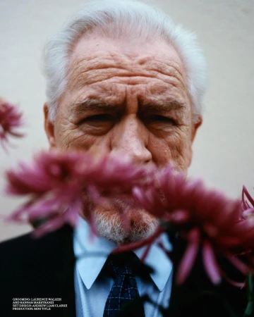 Portrait of an elderly adult man with white hair, wearing a suit and tie, partially obscured by pink flowers against a plain background.