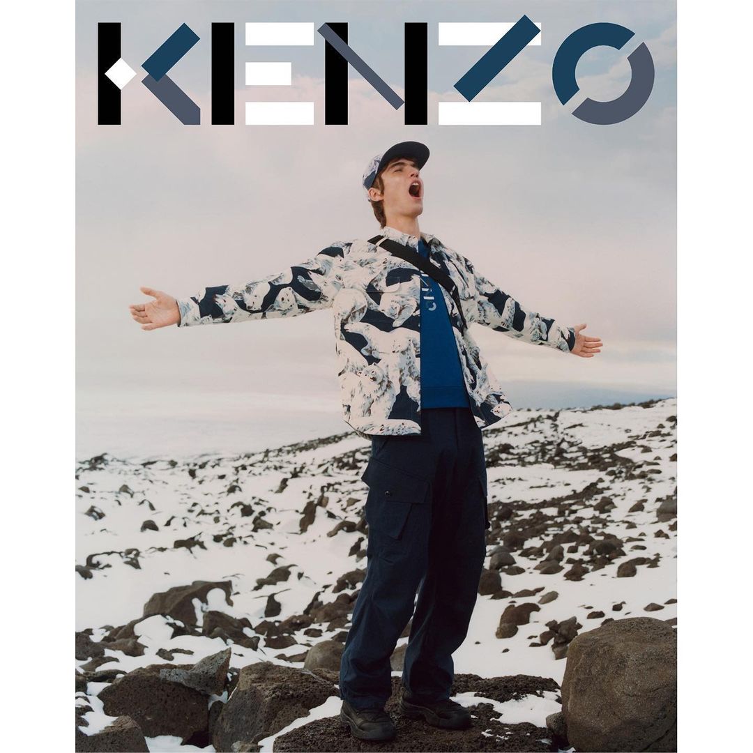 Man stands in snowy landscape wearing a jacket and cap with arms outstretched. The KENZO logo is in the upper portion of the image.