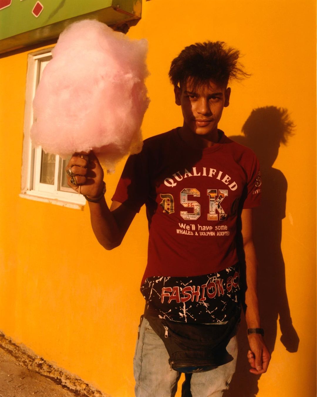 Young man holds large pink cotton candy against a vibrant yellow wall with a small window; outdoor setting with casual fashion.
