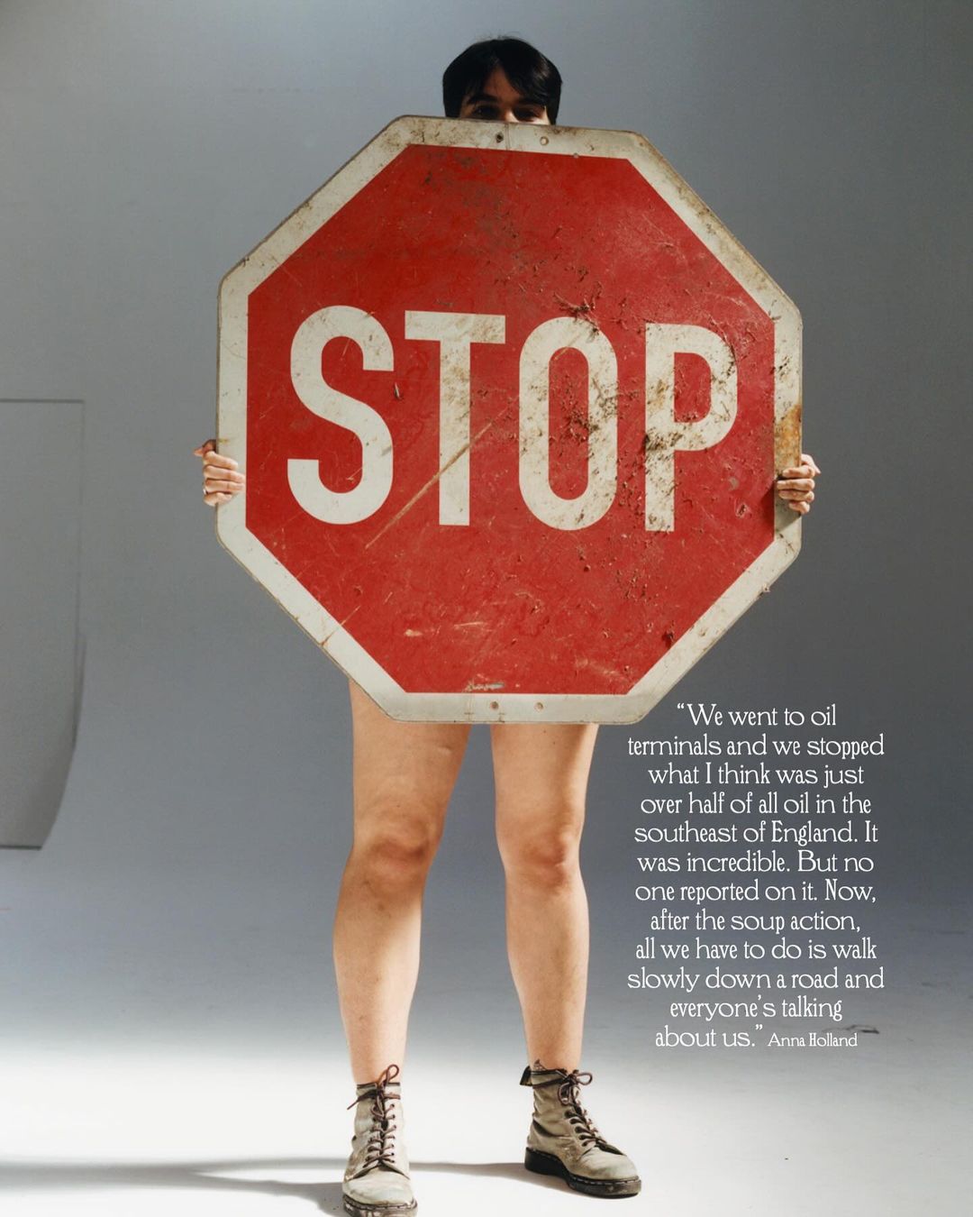 Person standing behind a red and white stop sign, wearing boots, against a grey backdrop in a studio setting.