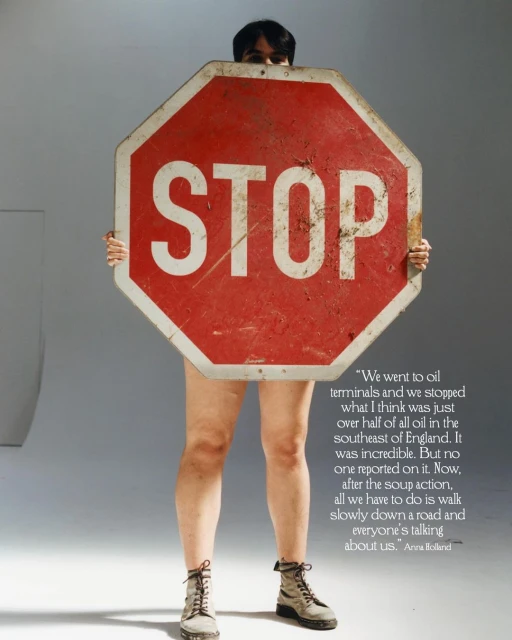 Person standing behind a red and white stop sign, wearing boots, against a grey backdrop in a studio setting.