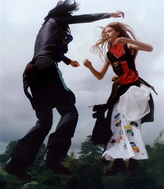 Two young adults in colorful streetwear jump outdoors on a cloudy day above green treetops; one in blue, one in white and red.
