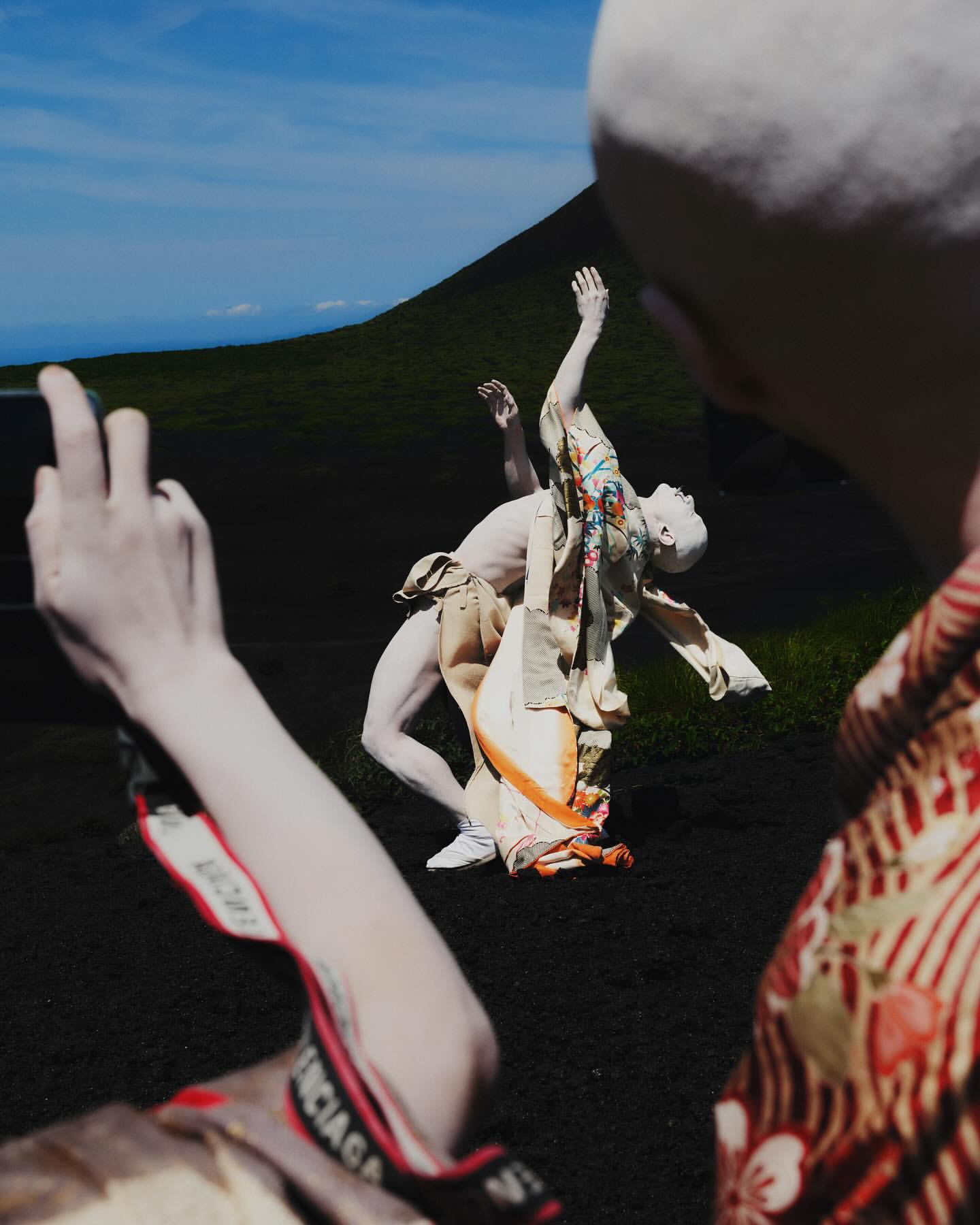 Two pale-skinned figures in loose garments pose outdoors near a dark hill under a bright sky, as a third person holds a phone.
