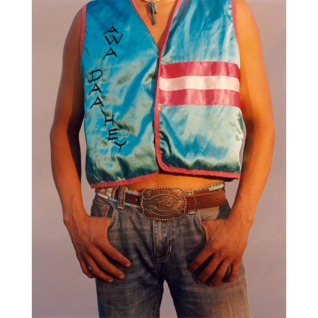 Torso of person wearing turquoise vest with pink stripes and belt buckle, against a neutral background, hands in pockets.