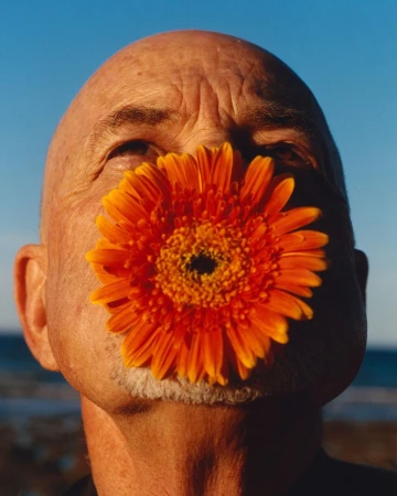 Close-up portrait of an elderly man holding a large bright orange gerbera daisy in front of his mouth against a blue sky.