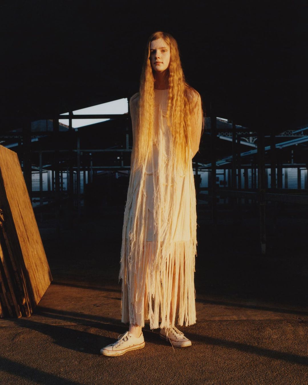 A young woman with long hair stands in a fringed dress and sneakers. She is posing in front of a dark industrial space.