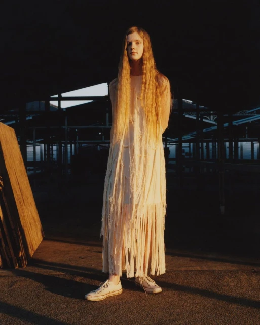 A young woman with long hair stands in a fringed dress and sneakers. She is posing in front of a dark industrial space.