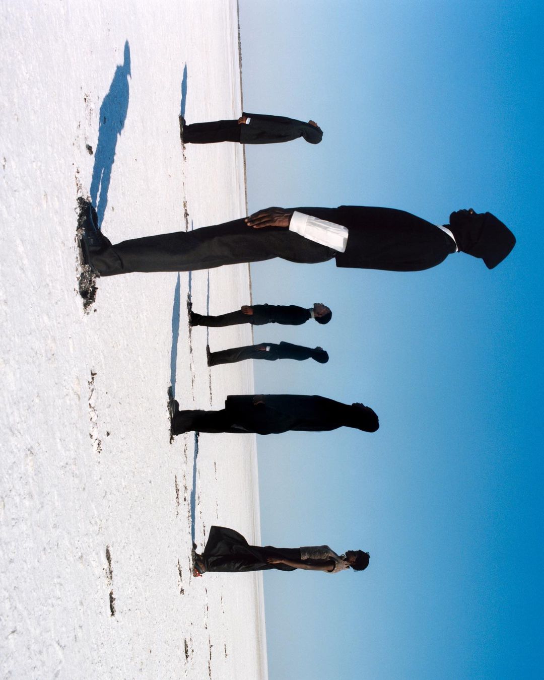Five men in suits stand in a line on a white surface under a blue sky; a creative perspective that plays with scale and landscape.