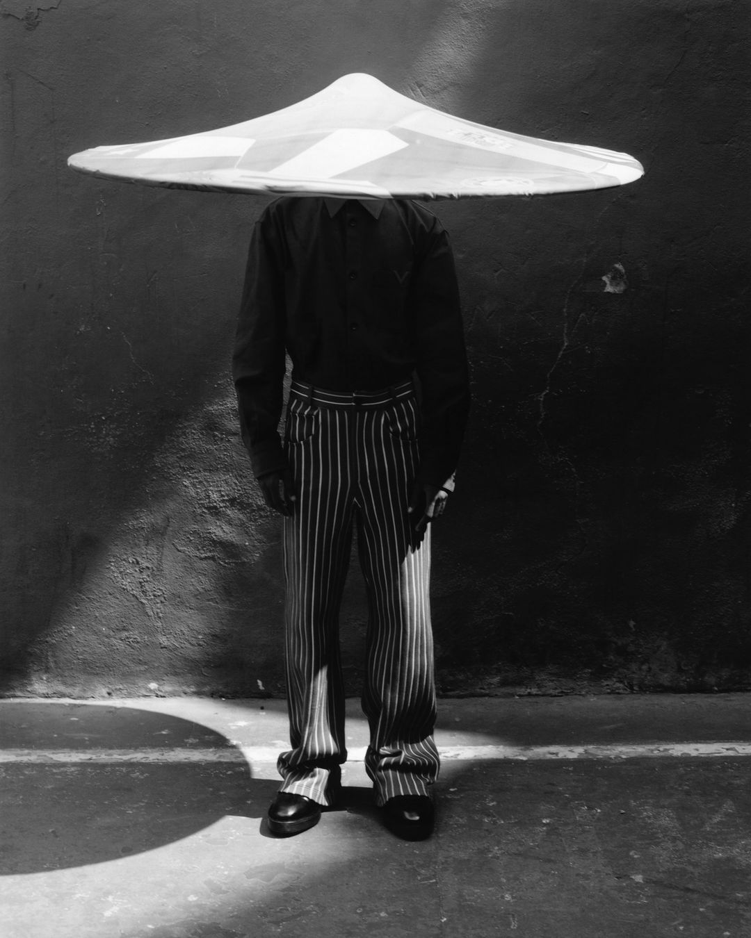 Monochrome image of a person wearing a large conical hat, pinstripe pants, and dark shirt, standing against a textured wall with shadows.