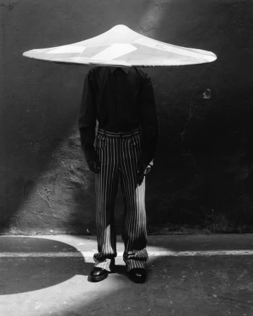 Monochrome image of a person wearing a large conical hat, pinstripe pants, and dark shirt, standing against a textured wall with shadows.