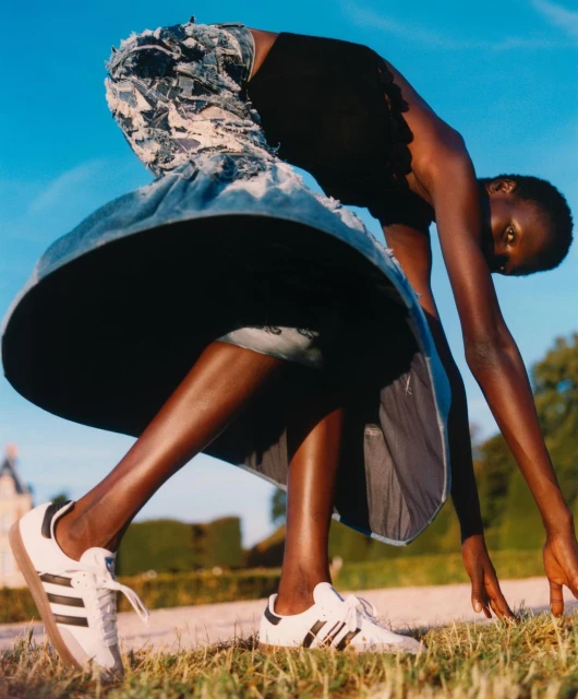 Eye-level outdoor fashion shot of a slender Black woman posing with white sneakers, a black top, and a blue denim skirt against a clear blue sky.