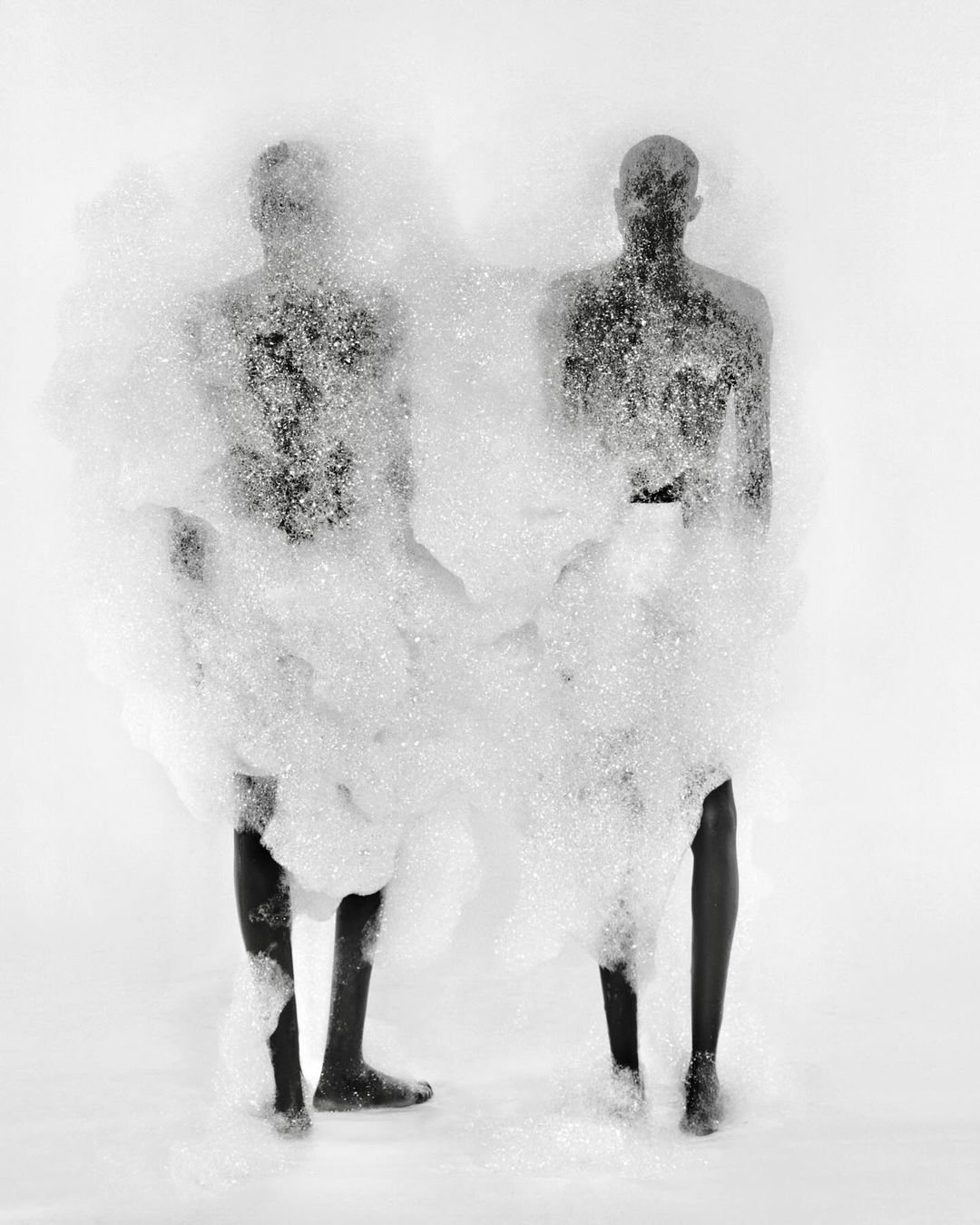 Two figures are nearly obscured by a cloud of white foam in a black-and-white studio photograph with a bright white background.