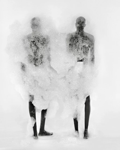 Two figures are nearly obscured by a cloud of white foam in a black-and-white studio photograph with a bright white background.