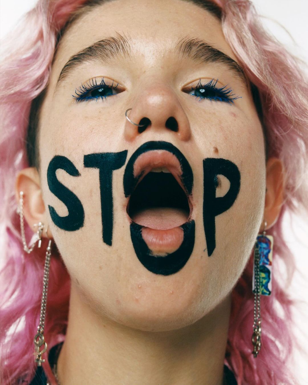 Eye-level close-up of a young woman with pink hair and a piercing, with the word 'STOP' written over her mouth and chin.