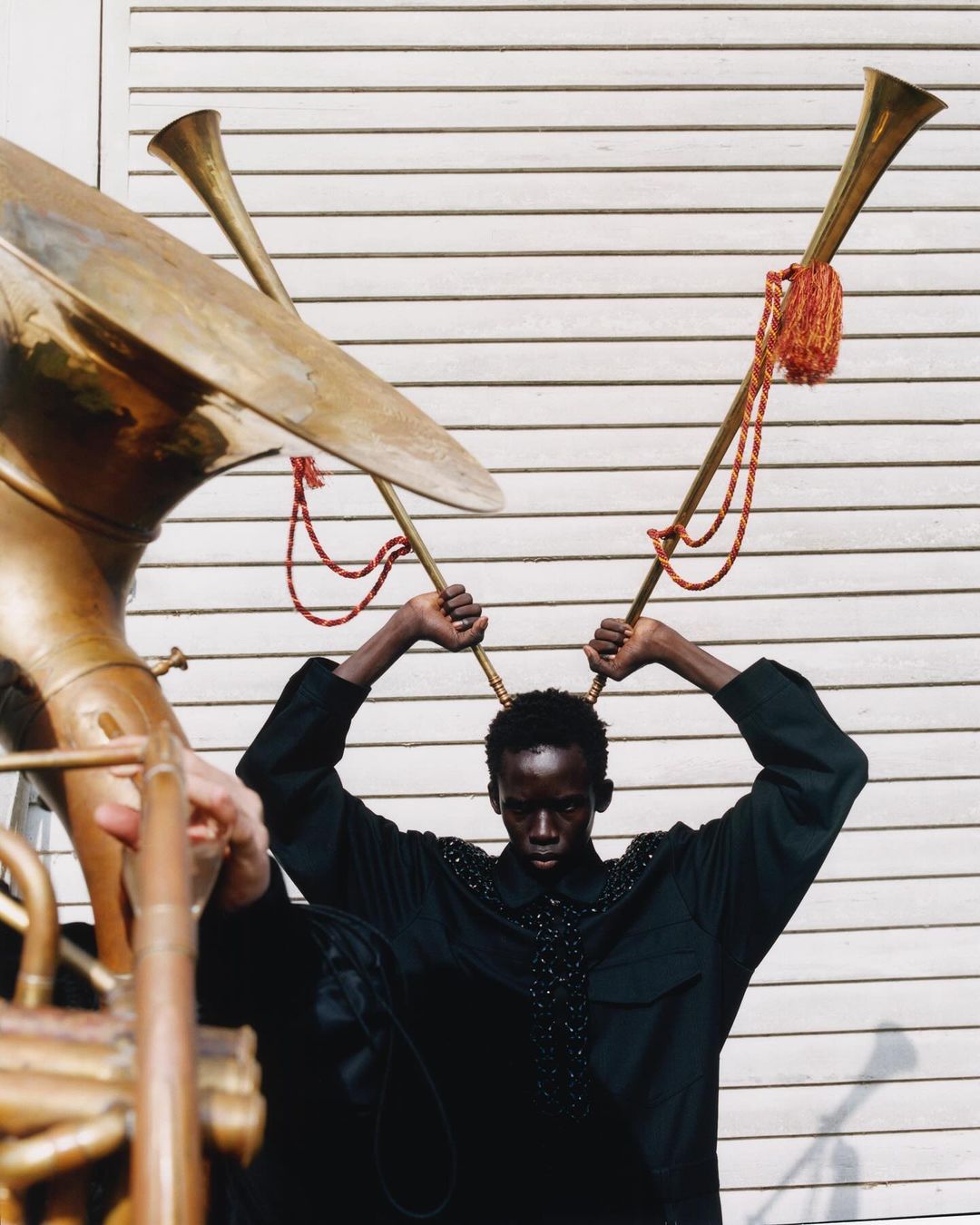 Fashion shot of a model in black shirt posing with two large brass horns against a white wall, creating a surreal and musical aesthetic.