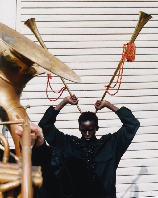 Fashion shot of a model in black shirt posing with two large brass horns against a white wall, creating a surreal and musical aesthetic.