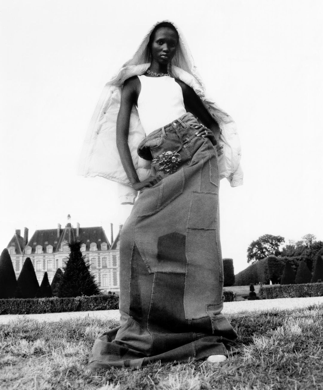 Fashion shot of an adult woman modeling a patchwork denim maxi skirt and white top, posing in a park with a chateau in the background.