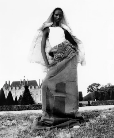 Fashion shot of an adult woman modeling a patchwork denim maxi skirt and white top, posing in a park with a chateau in the background.