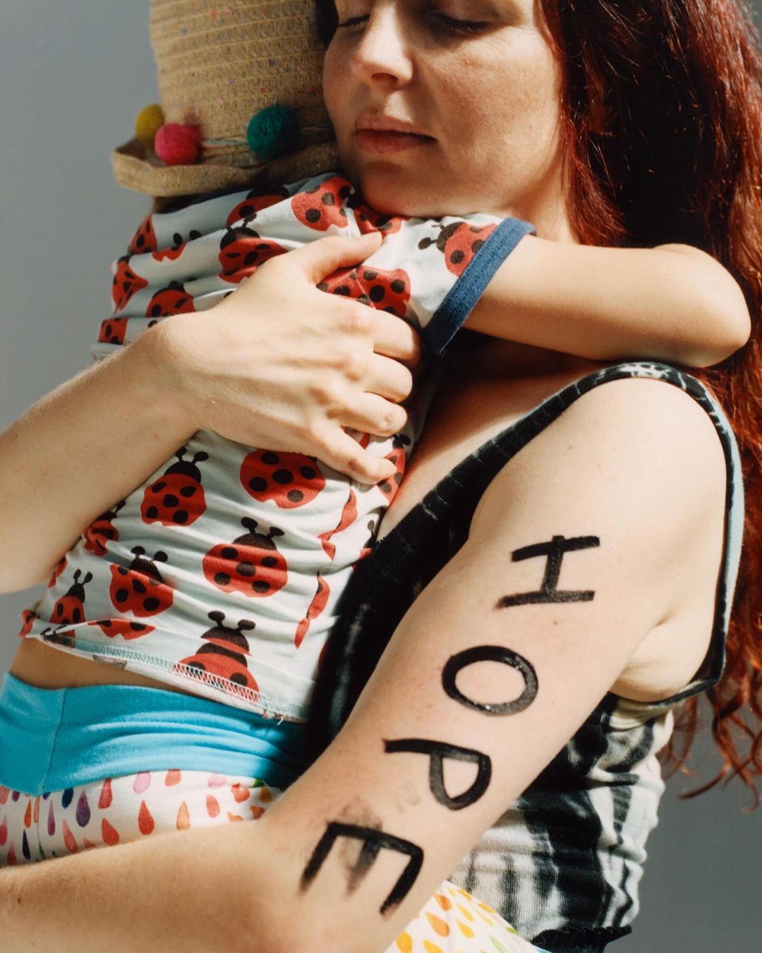 A mother with red hair holds her young child wearing a ladybug shirt and straw hat; the word 'HOPE' is painted on the mother's arm.