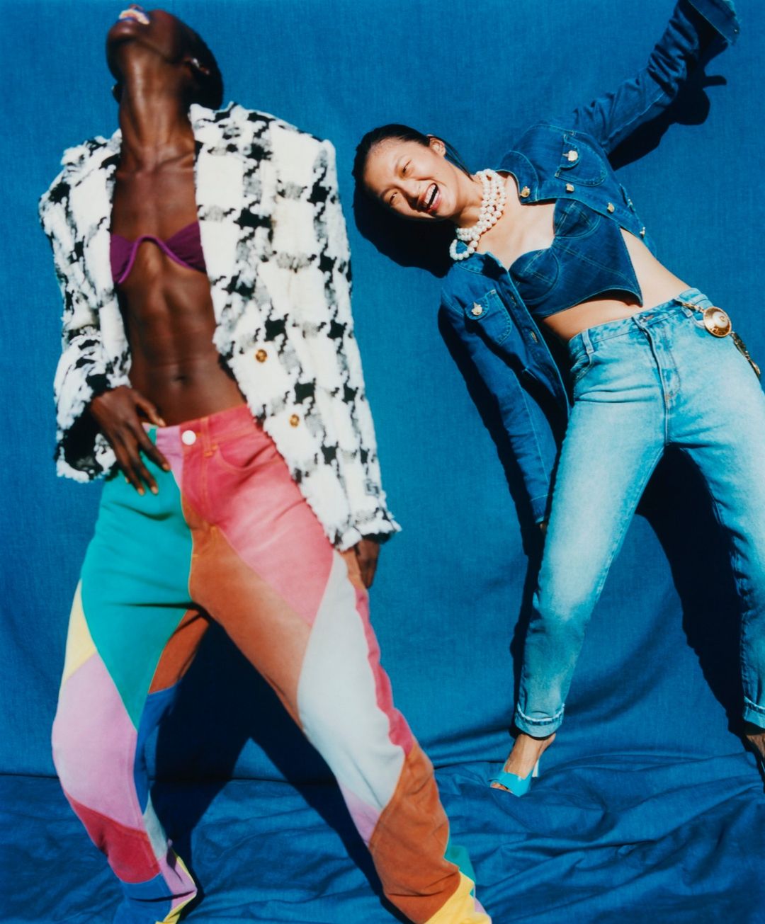 Two models pose in colorful retro fashion against a blue backdrop. One wears a textured jacket and patchwork pants, the other in denim.