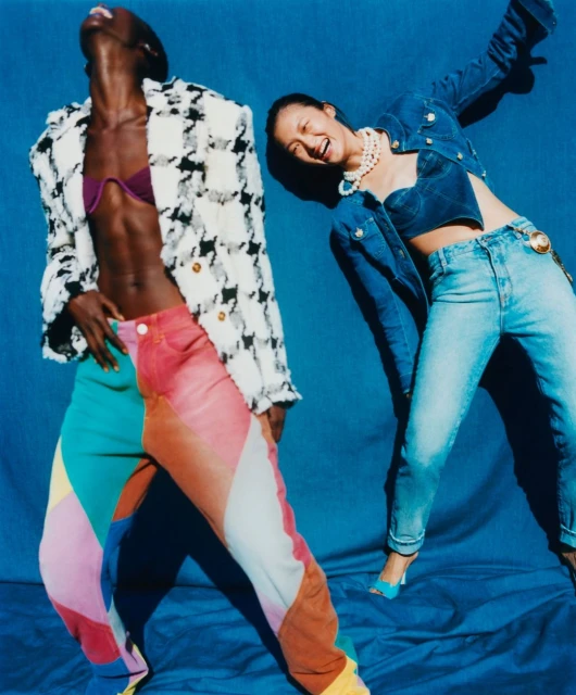 Two models pose in colorful retro fashion against a blue backdrop. One wears a textured jacket and patchwork pants, the other in denim.