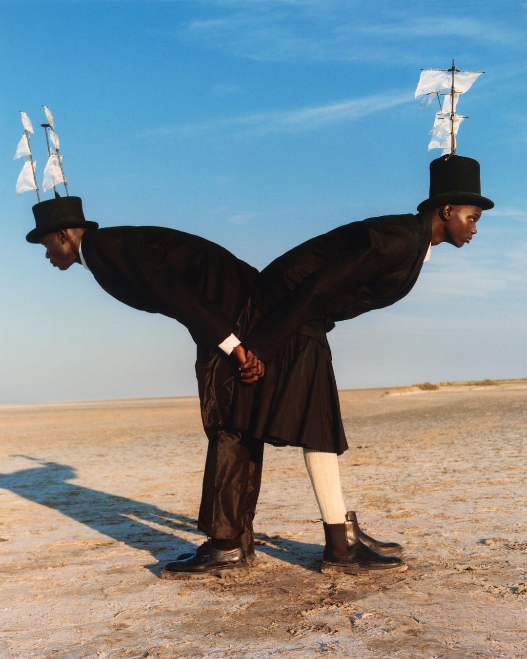 Two figures in surreal fashion, posed against a sky and beach, with ship models atop hats, creating a striking visual statement.