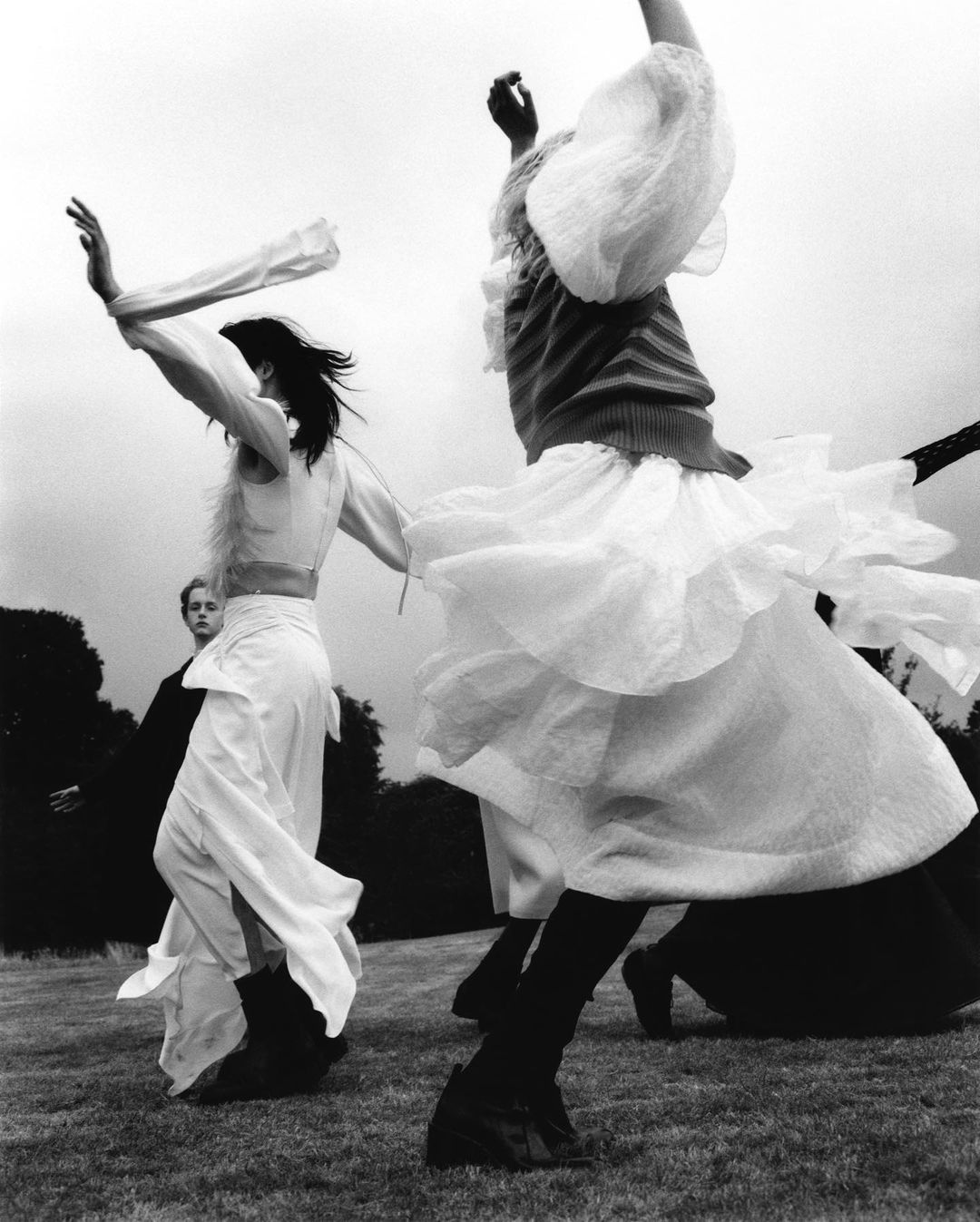 Three women in flowing white dresses dance in an open grassy field under an overcast sky, captured in a vintage black and white style.