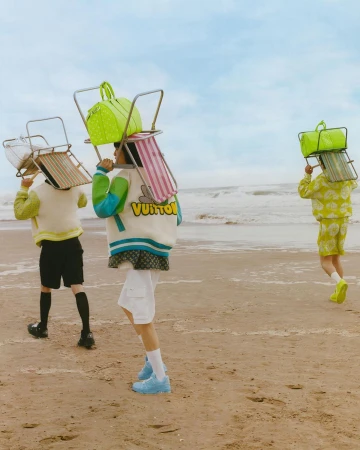 Three figures in streetwear carry beach chairs and bright bags on a beach with a blue sky and water, conveying a summery, seaside mood.