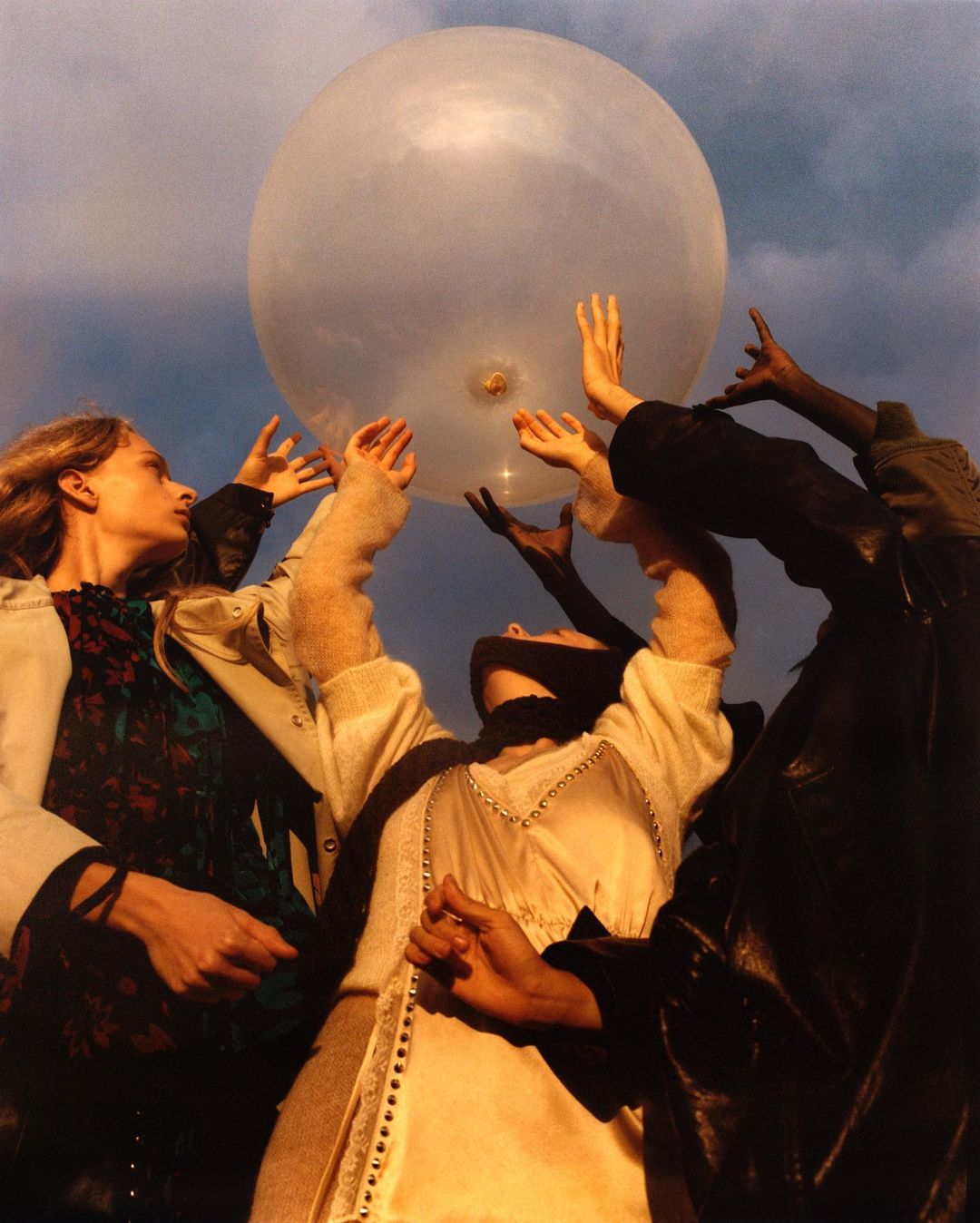 Group of people touching a large, transparent balloon in an outdoor setting with dramatic lighting and sky in the background.