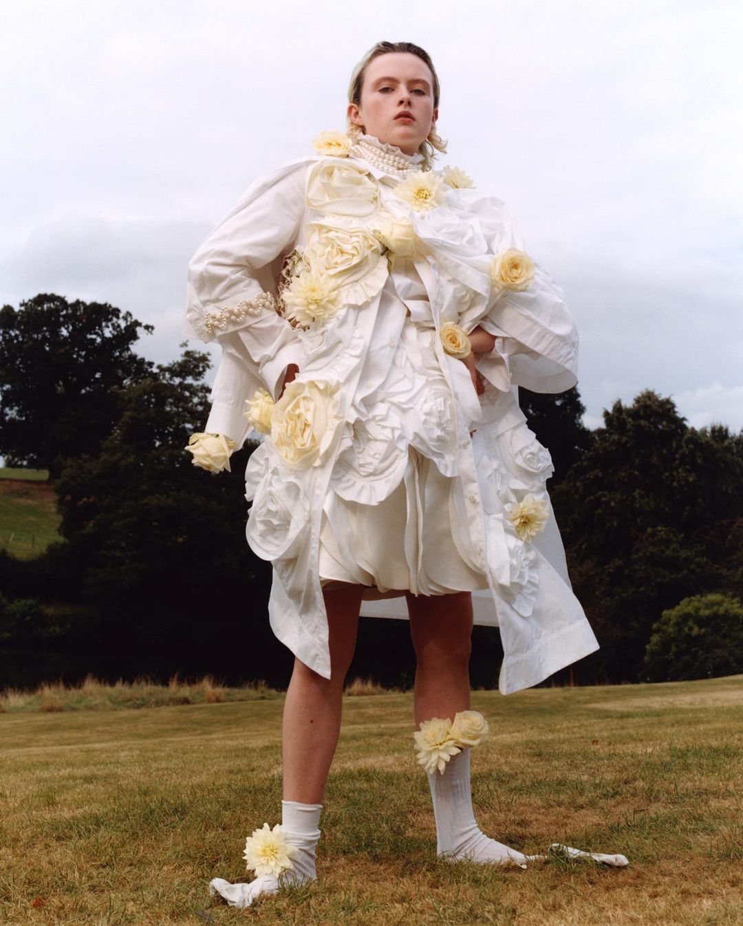 An adult posing outdoors wearing a dramatic white garment with pale yellow flower appliques on the bodice, short skirt, and socks. Cloudy sky.