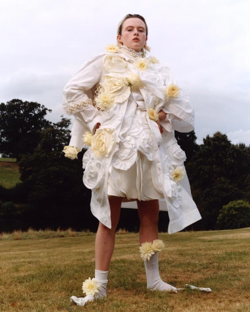 An adult posing outdoors wearing a dramatic white garment with pale yellow flower appliques on the bodice, short skirt, and socks. Cloudy sky.