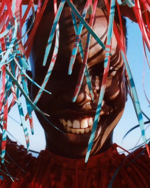 Close-up studio shot of a smiling Black woman with light blue and red zip ties creating a hair covering against a bright blue sky backdrop.