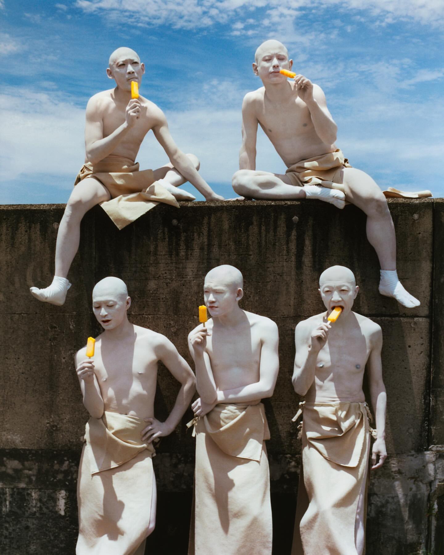 Five people with white skin eat orange popsicles outdoors against a blue sky. Three stand, two sit above on a concrete structure.