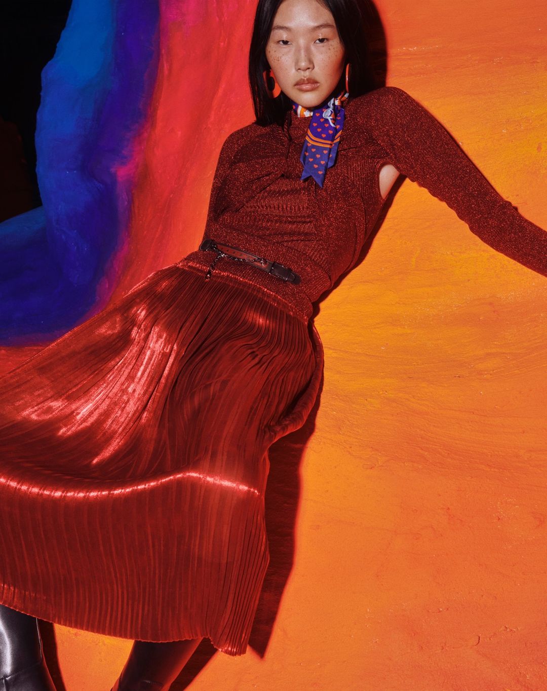 Fashion shot featuring an Asian woman in a metallic brown top and red pleated skirt against a colorful background in a photo studio.